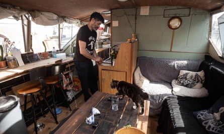مردی در اتاق نشیمن / آشپزخانه قایق خانه اش.  گربه ای روی میز قهوه راه می رود.